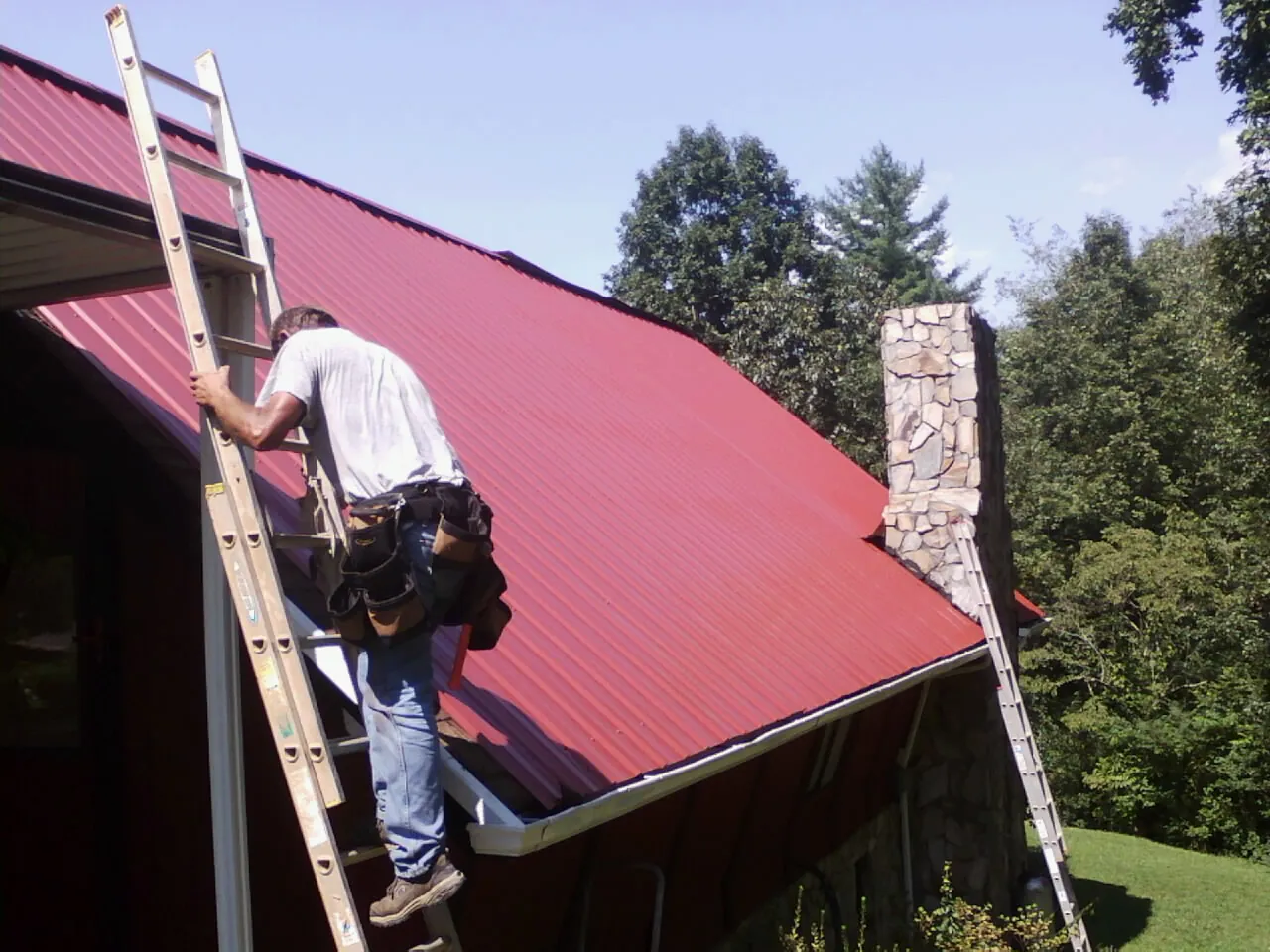 Roofer on a ladder beside a red metal roof and stone chimney — Soffit & Fascia Repair by Roofing Brothers in Sayre